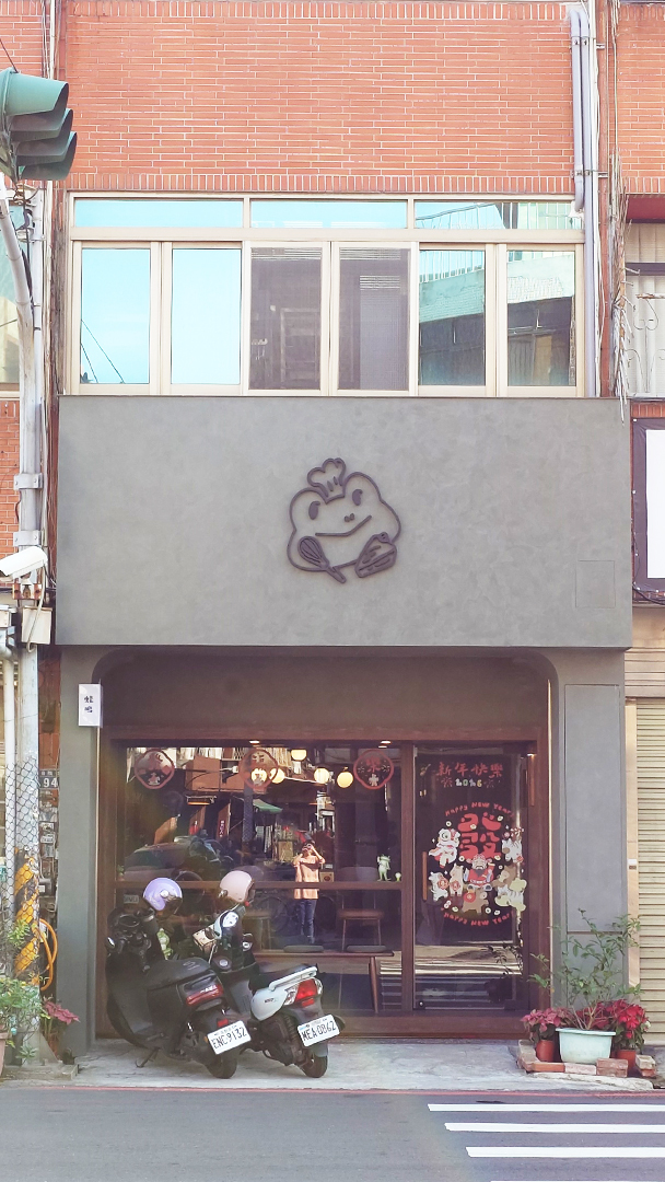 蛙鳴甜點外觀，灰綠色外牆上，有隻青蛙拿著烘焙工具的logo